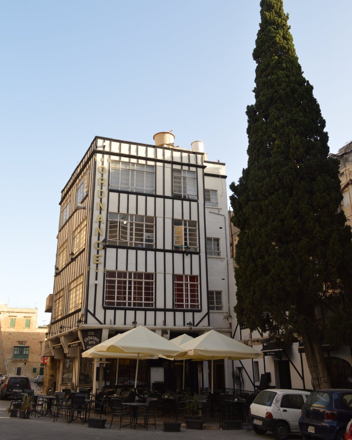 The black-and-white timbered facade of The Ordnance on its square in Valletta, cream umbrellas open below and a tall cypress alongside
