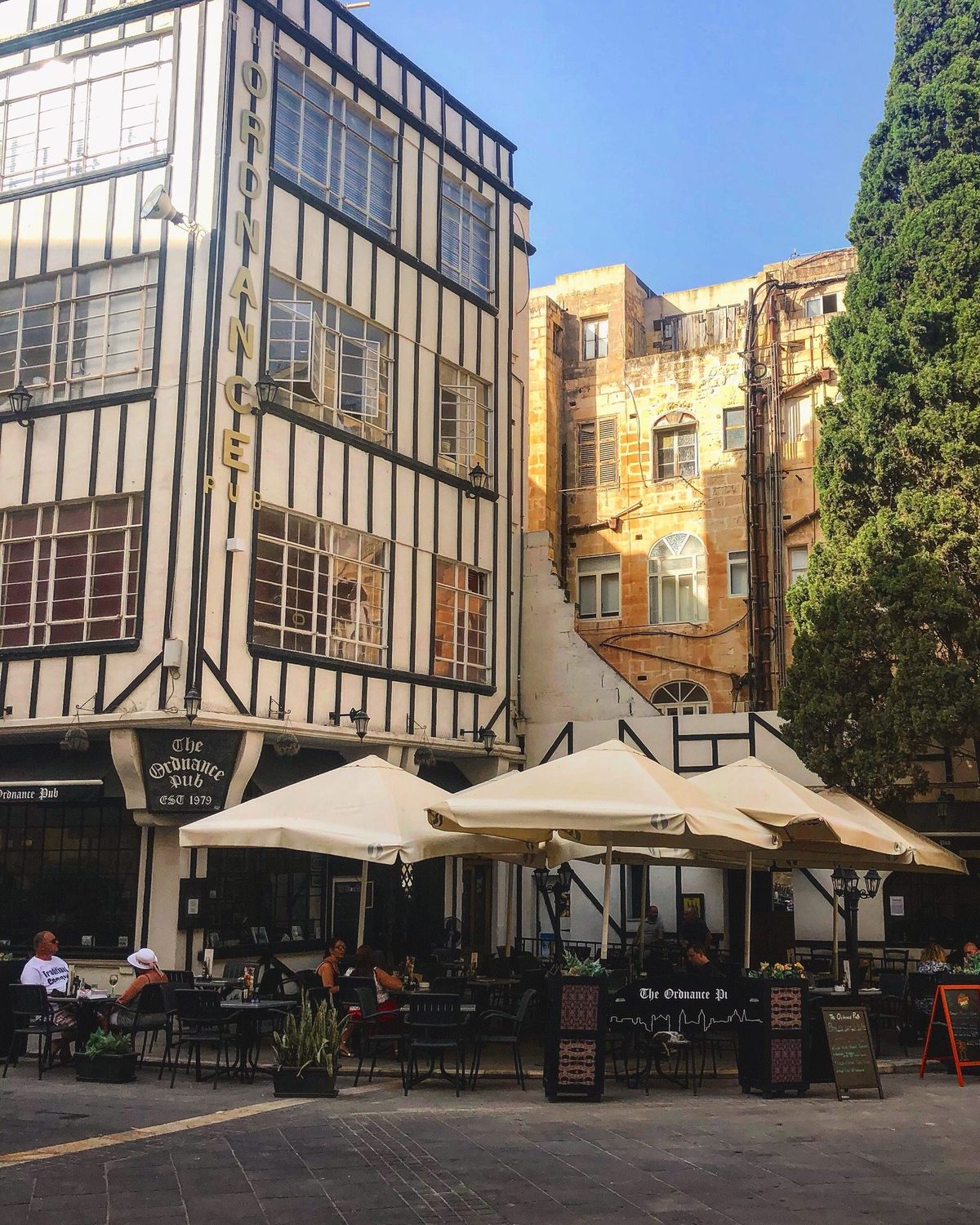 The Ordnance on its square in daylight, cream umbrellas open over the tables